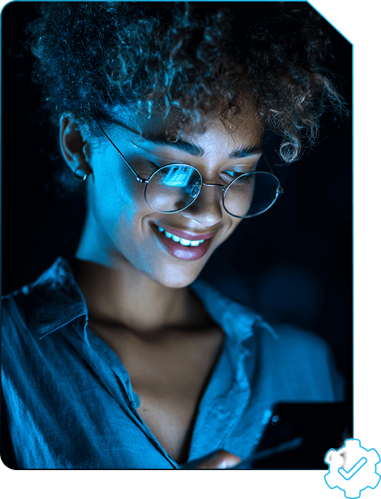 Mulher negra de cabelo cacheado usando óculos redondo e camiseta azul, em ambiente escuro e com luz azul, olhando para o celular e sorrindo